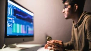 Homem segura moeda de bitcoin em frente ao computador com uma tela de investimentos aberta